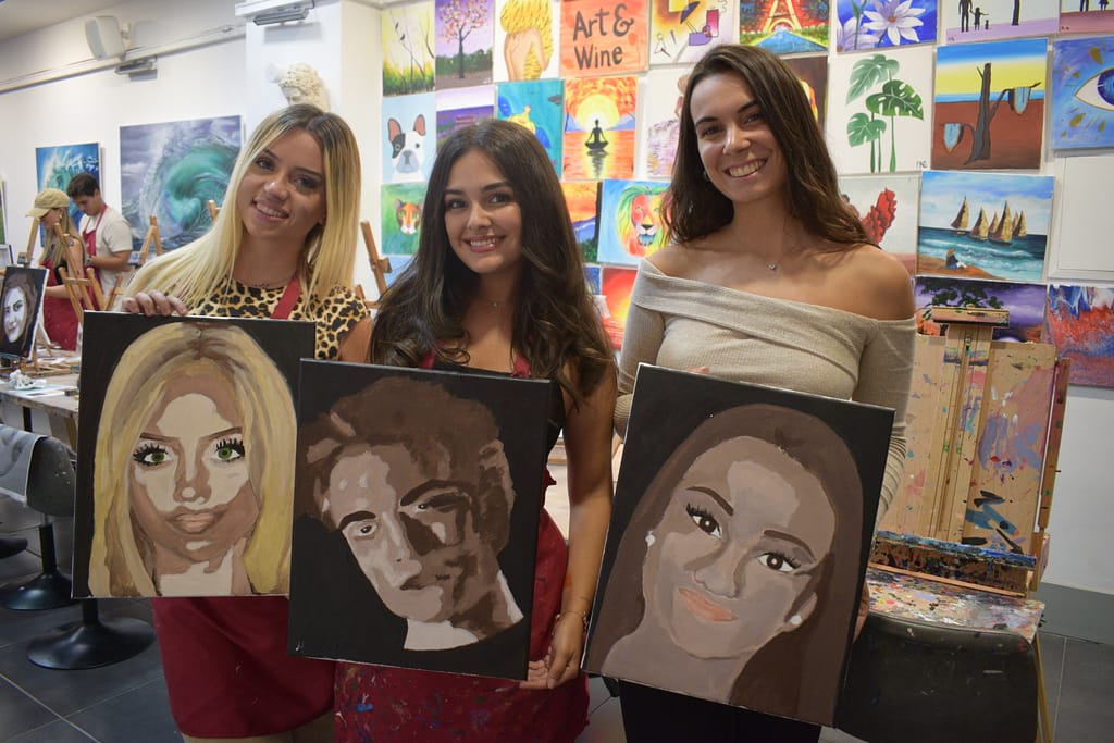 Tres mujeres en un taller de pintura sostienen sus lienzos, mostrando retratos pintados de rostros en tonos marrones y beige. Están de pie juntas, sonriendo, mientras posan con sus creaciones artísticas en un entorno lleno de coloridos cuadros colgados en la pared